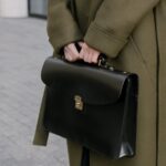 View of a person's midsection only, wearing a long green coat and holding a black leather briefcase, standing outside on grey cobblestone.